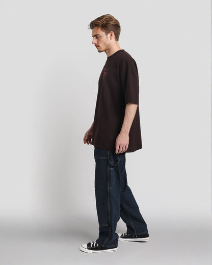 Man wearing a brown t-shirt and dark jeans standing against a plain background